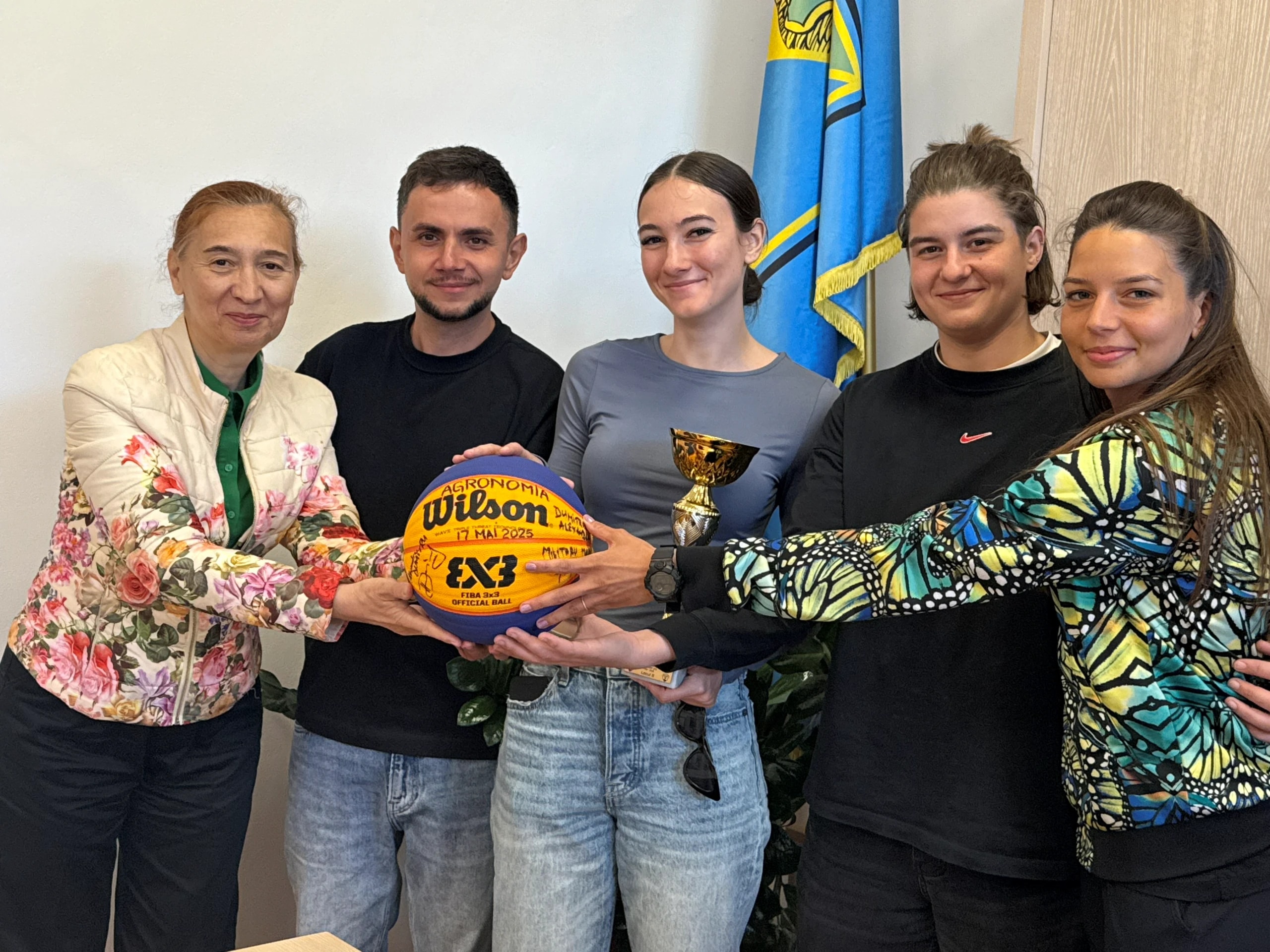 Mingea de 3x3 semnată și trofeul, ținute împreună de sportive și de oameni ai clubului