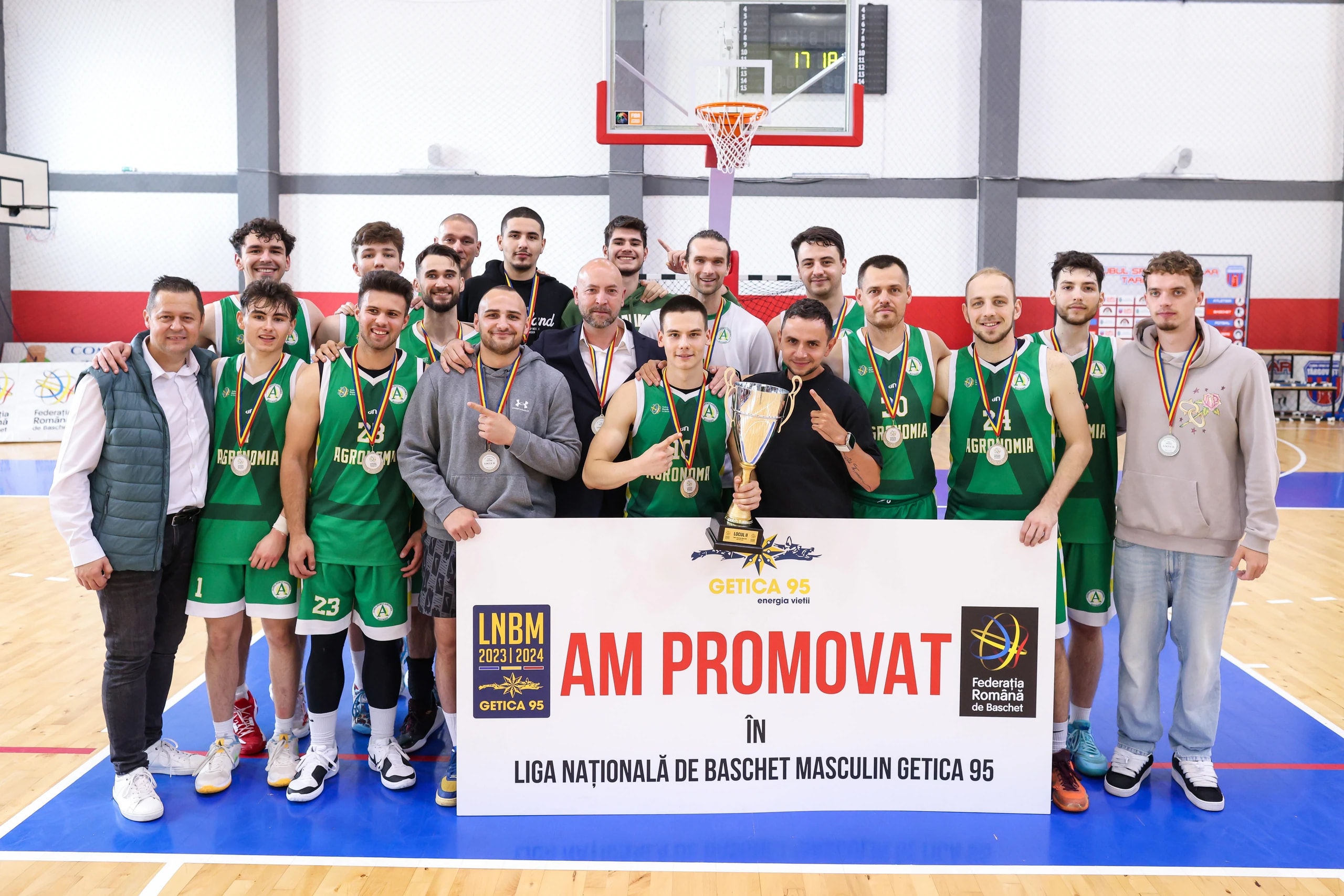 Echipa masculină de baschet, cu medaliile și cupa, în fața bannerului de promovare în Liga Națională